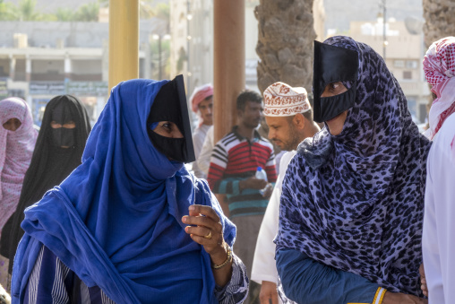 Two Omani women wearing colorful headscarves and black face masks conversing on a street with men nearby, Ad Dakhiliyah Region, Nizwa, Oman