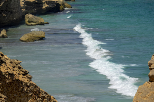 Close-up of a wave breaking in turquoise water between two limestone cliff outcrops with birds flying above, Dhofar Governorate, Taqah, Oman