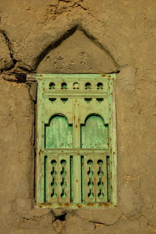 Close-up of a weathered green wooden window with a carved lattice shutter set into a cracked mudbrick wall, Dhofar Governorate, Mirbat, Oman