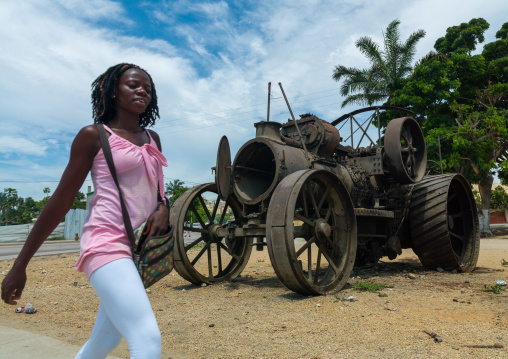 Angolan woman walking in front of antique steam traction engines displayed along the road, Benguela Province, Benguela, Angola