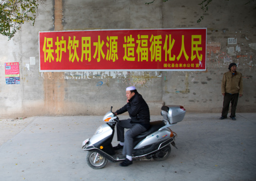 Salar ethnic minority man on a scooter passing in front of a chinese propaganda poster about saving water, Qinghai province, Xunhua, China