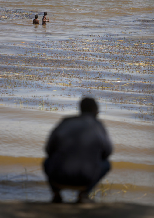 Silhouette of a man squatting by zway lake while kids are swimming, Zway, Ethiopia