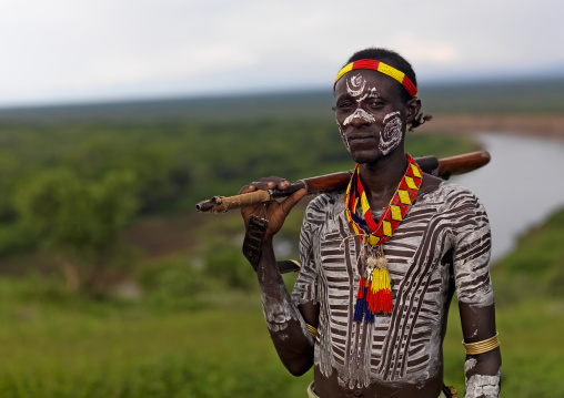 Portrait Of A Karo Tribe Man With Traditional Body Paint Holding A Kalashnikov And Posing Over The Omo River, Korcho Village, Omo Valley, Ethiopia
