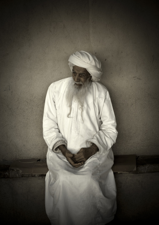 Old Omani Man In White Dishdasha Sitting In Front Of The Wall, Nizwa, Oman