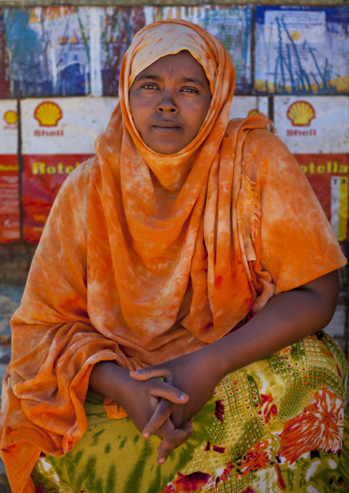 A Mature Woman Wearing An Orange Veil Sitting On The Ground With A SHell Board In The Background, Lasadacwo Village, Somaliland