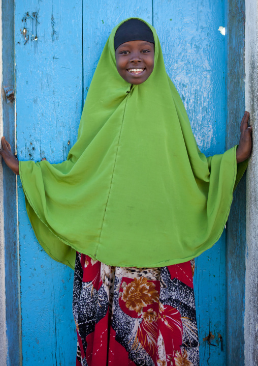Portrait Of A Little Girl Wearing A Green Hijab And A Red Patterned Dress Leaning On A Blue Painted Wooden Door, Boorama, Somaliland