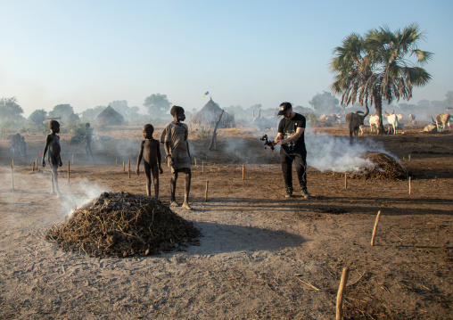 Tourist filming Mundari tribe boy taking care of the bonfires made with dried cow dungs, Central Equatoria, Terekeka, South Sudan