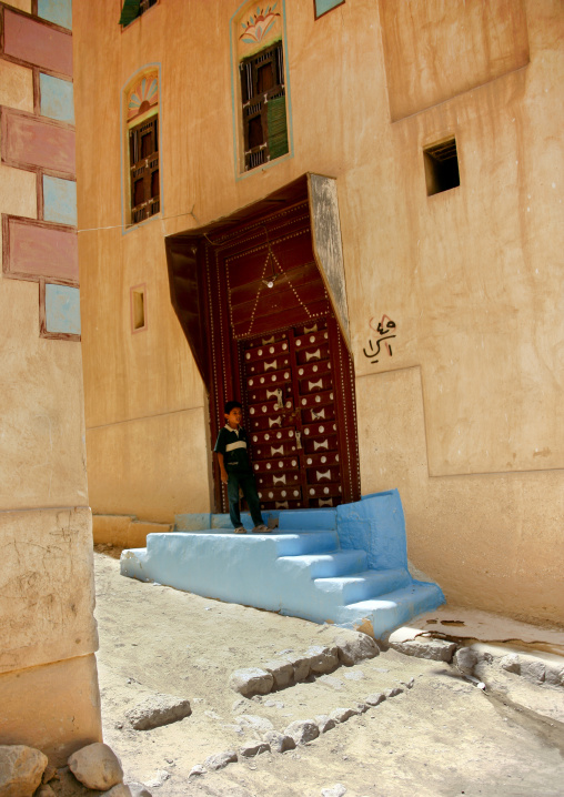 Boy Standing On The Blue Doorstep Of The Wooden Carved Door Of A Typically Painted Building, Wadi Doan, Yemen