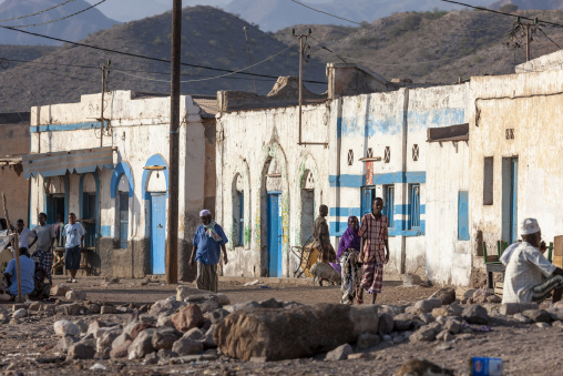 Local residents walk through a street lined with blue-trimmed colonial buildings beneath rugged mountains, Tadjourah Region, Tadjoura, Djibouti