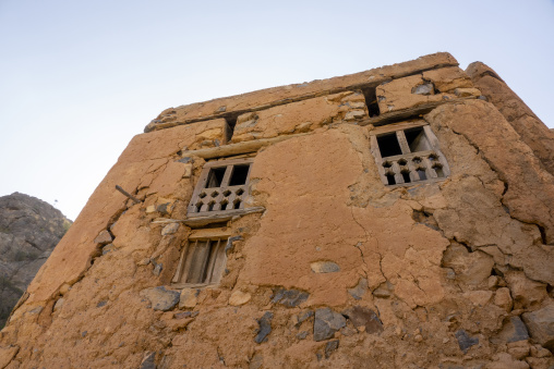 Cracked mudbrick facade of an abandoned house with carved wooden window screens seen from a low angle, Ad Dakhiliyah Region, Al Hamra, Oman