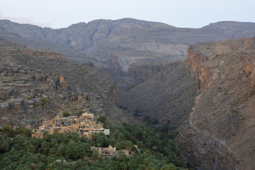 Village of Misfat al Abriyyin nestled among palm trees at the entrance to a dramatic rocky canyon in hazy morning light, Ad Dakhiliyah Region, Misfat al Abriyyin, Oman