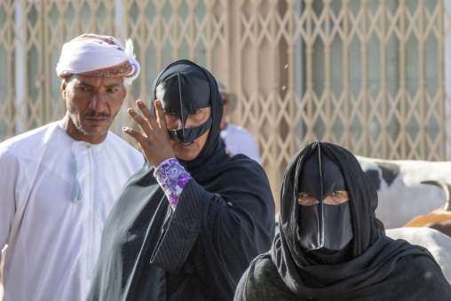 Omani man in a white turban standing near two women wearing traditional black face masks at the Nizwa livestock market, Ad Dakhiliyah Region, Nizwa, Oman