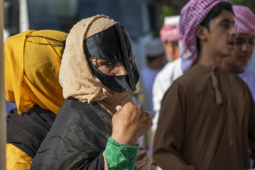 Omani woman adjusting her beige headscarf and black leather face mask amid a crowd at the Nizwa livestock market, Ad Dakhiliyah Region, Nizwa, Oman