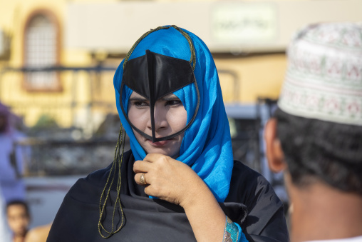 Omani woman tying her blue headscarf trimmed with gold cord over a black leather face mask, Ad Dakhiliyah Region, Nizwa, Oman