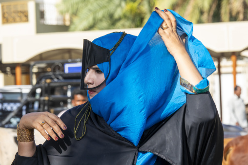 Omani woman raising her blue headscarf over a black leather face mask while wearing gold rings and bracelets, Ad Dakhiliyah Region, Nizwa, Oman