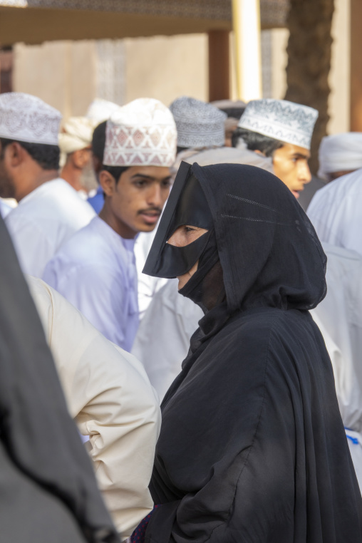 Omani woman in a black abaya and leather face mask walking through a crowd of men wearing white dishdasha and embroidered caps, Ad Dakhiliyah Region, Nizwa, Oman