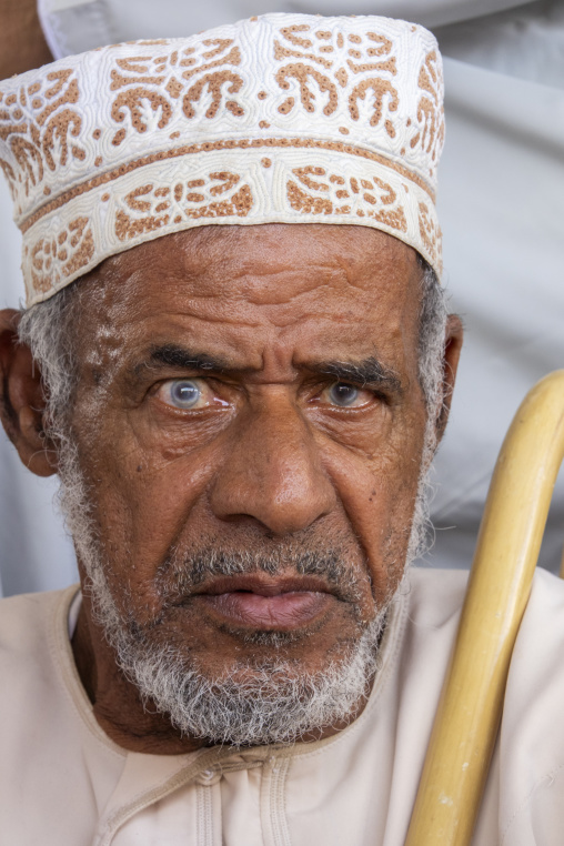 Close-up portrait of an elderly man wearing an embroidered kummah cap and holding a wooden cane, Ad Dakhiliyah Region, Nizwa, Oman