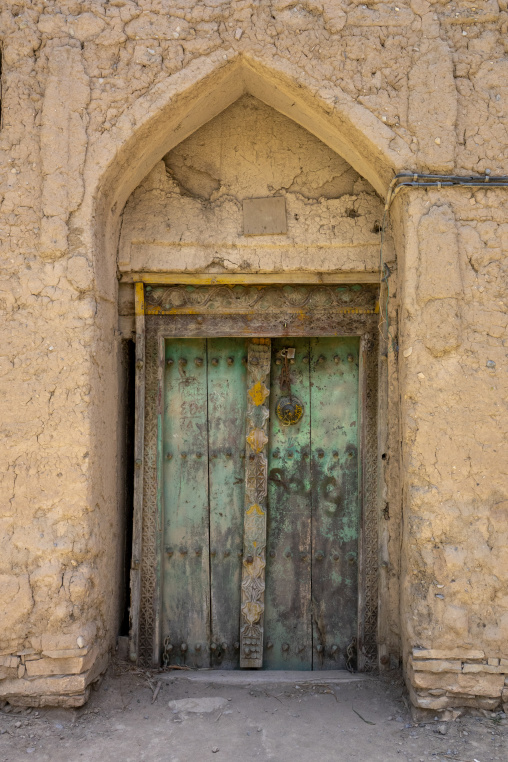 Weathered teal painted wooden door with a carved decorative panel set in a mudbrick archway, Ad Dakhiliyah Governorate, Birkat Al Mouz, Oman