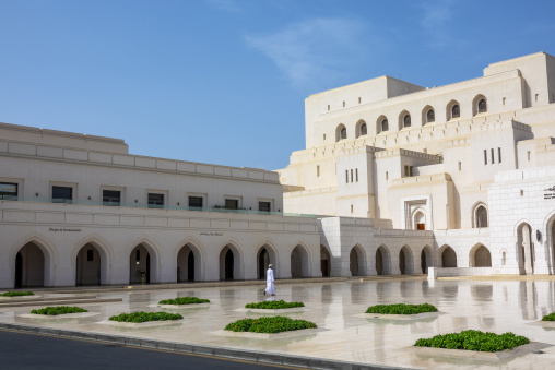 Man in a white dishdasha walking across a wet reflective marble courtyard in front of the Royal Opera House Muscat and its shops and restaurants wing under a blue sky, Governorate of Muscat, 