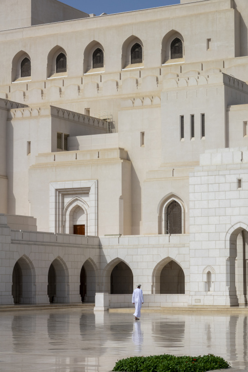 Man in a white dishdasha and cap walking beneath the arched colonnade of the Royal Opera House Muscat facade with latticed windows above, Governorate of Muscat, Muscat, Oman