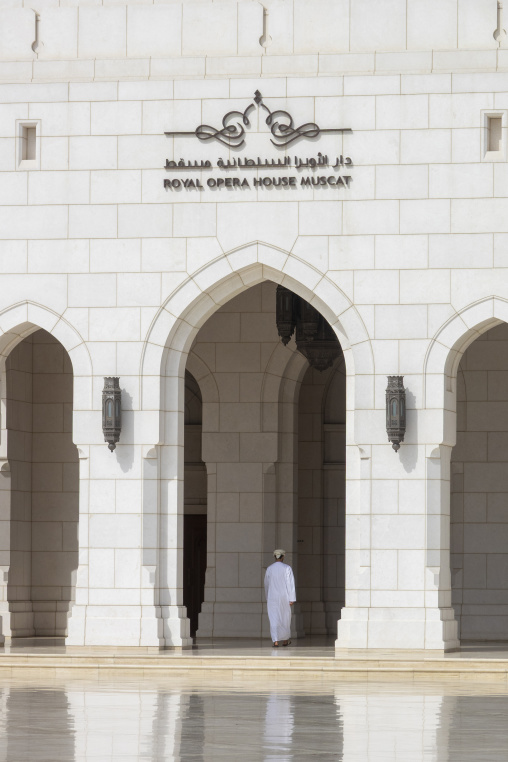 Man in a white dishdasha walking through the arched entrance below the bilingual Royal Opera House Muscat sign flanked by ornate lanterns, Governorate of Muscat, Muscat, Oman