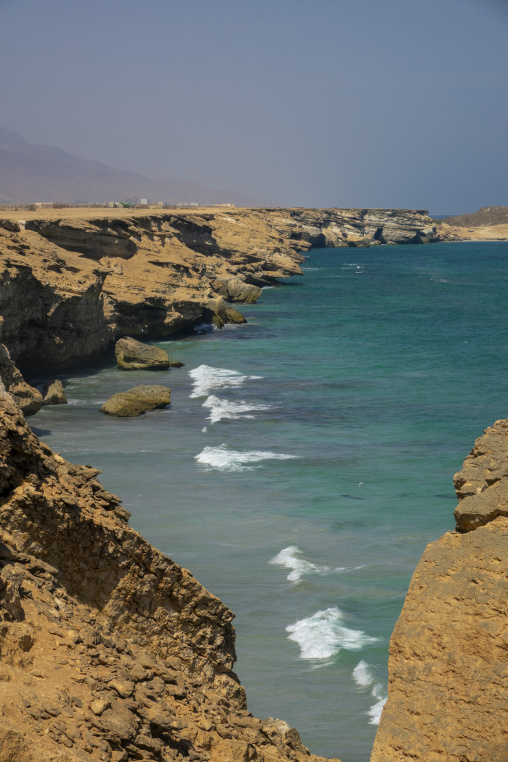 Rugged limestone cliffs overlooking a turquoise sea with waves breaking below and a hazy coastal settlement in the distance, Dhofar Governorate, Taqah, Oman