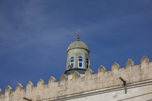 Tiled minaret dome topped with a crescent moon finial rising above a crenellated mosque wall against a clear blue sky, Dhofar Governorate, Wadi Dirba, Oman