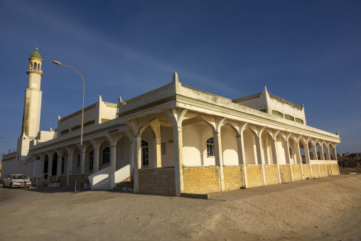 Old mosque with a green onion-domed minaret and an arched cream colonnade beside a sandy street, Dhofar Governorate, Mirbat, Oman