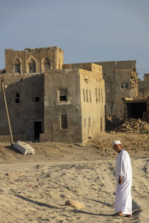 Smiling man in a white dishdasha and cap walking past a cracked crumbling mudbrick building with an overturned boat nearby, Dhofar Governorate, Mirbat, Oman