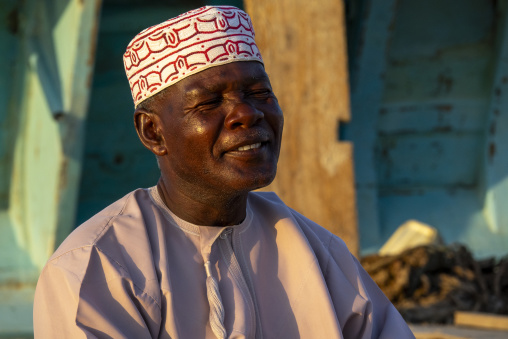 Portrait of a smiling man wearing an embroidered Omani cap and grey dishdasha squinting in the sunlight, Dhofar Governorate, Mirbat, Oman