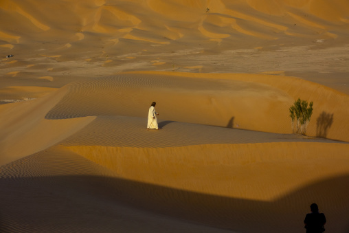 Omani man in a white robe walking with a wooden stick across sand dunes, Dhofar Governorate, Rub al Khali, Oman