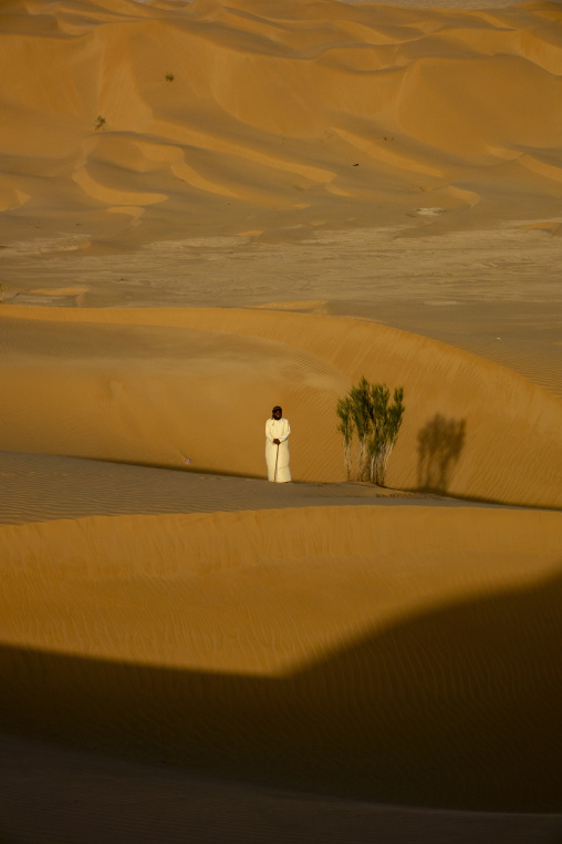 Man in a white robe standing beside a green desert shrub on a sand dune, Dhofar Governorate, Rub al Khali, Oman